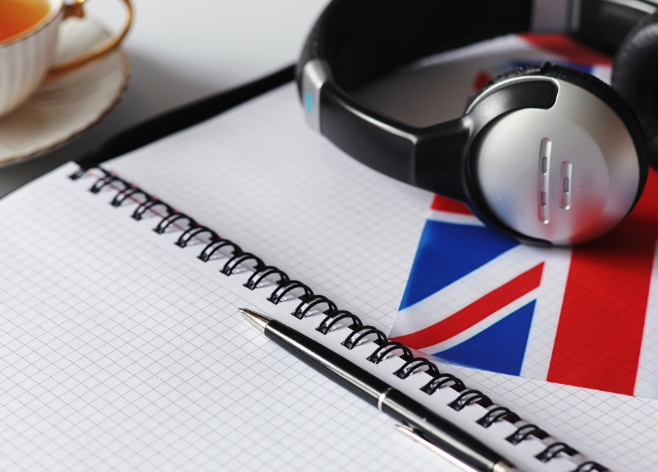 Otwarty duży zeszyt na spirali, na zeszycie leżą słuchawki i długopis, pod słuchawkami flaga Wielkiej Brytani.