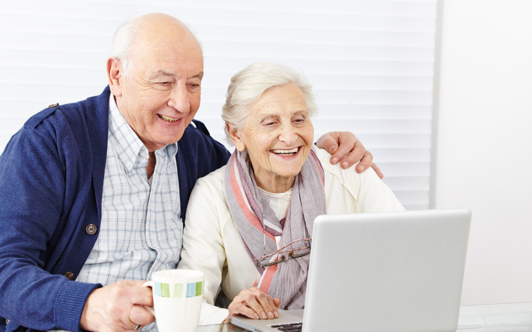 Siwowłosa starsza kobieta, siedzi przed laptopem, uśmiecha się szeroko. Obok niej siedzi starszy siwy mężczyzna obejmuje ją ramieniem.